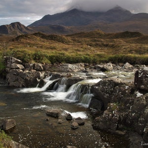 May include: A cascading waterfall flows over moss-covered rocks in a rugged, mountainous landscape. The water is clear and white, and the rocks are dark gray. The mountains in the background are covered in green vegetation.