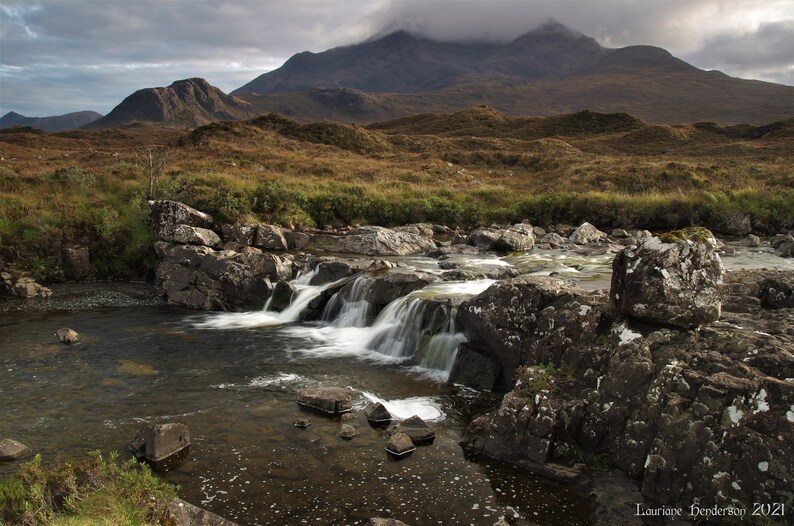 May include: A cascading waterfall flows over moss-covered rocks in a rugged, mountainous landscape. The water is clear and white, and the rocks are dark gray. The mountains in the background are covered in green vegetation.