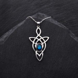 Puede incluir: Un colgante de nudo celta de plata con una incrustaci&oacute;n de concha de abul&oacute;n azul. El colgante cuelga de una cadena de plata sobre un fondo negro.