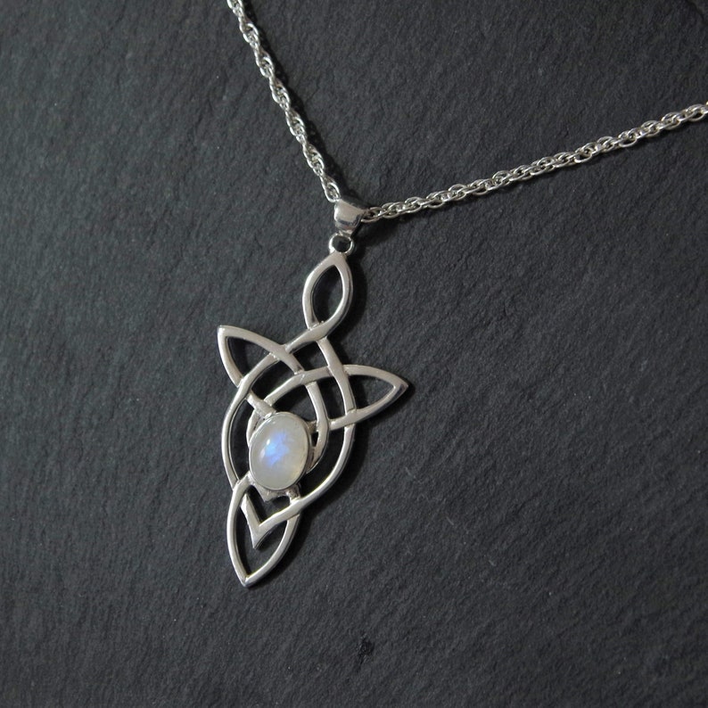 Puede incluir: Un collar de plata con un colgante de nudo celta con una piedra de luna en el centro. El collar est&aacute; sobre un fondo negro.