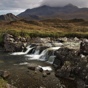 May include: A cascading waterfall flows over moss-covered rocks in a rugged, mountainous landscape. The water is clear and white, and the rocks are dark gray. The mountains in the background are covered in green vegetation.
