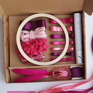 May include: A DIY dreamcatcher kit with a variety of pink and purple ribbons, feathers, and beads. The kit includes a wooden hoop, a box of supplies, and a completed dreamcatcher. The color palette is a mix of vibrant and soft pinks and purples.