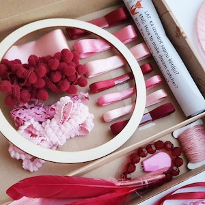 May include: A craft kit for making dream catchers, featuring a wooden hoop, ribbons, pom-poms, feathers, and beads in shades of pink and red. The kit includes a tube with text in English and Latvian.