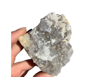 May include: A cluster of gray fluorite crystals with a cubic structure. The crystals are translucent and have a slight shimmer. The fluorite is a natural mineral and has a rough, uneven surface.