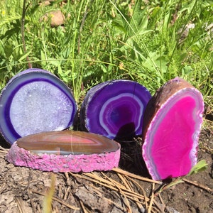 May include: Four agate slices with different patterns and colors. The slices are purple, pink, and brown with white and clear accents. The slices are arranged on a bed of pine needles.