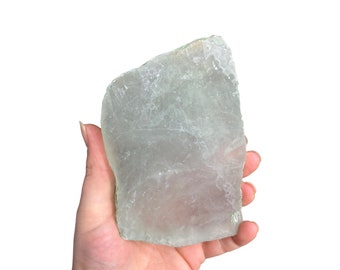 May include: A translucent, pale green crystal slab with irregular edges. The crystal is held in a hand against a white background, showcasing its natural texture and subtle color variations. The crystal appears to be a raw, unpolished gemstone.