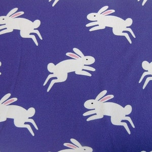 May include: A fabric with a repeating pattern of white rabbits with pink ears and black eyes, leaping across a solid purple background. The rabbits are stylized and cartoon-like, creating a playful and whimsical design.