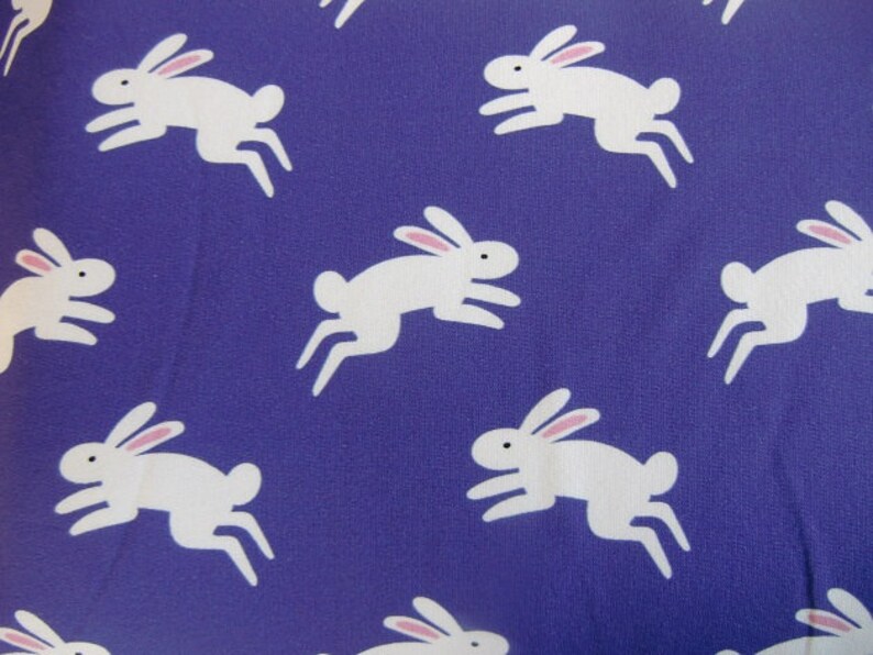 May include: A fabric with a repeating pattern of white rabbits with pink ears and black eyes, leaping across a solid purple background. The rabbits are stylized and cartoon-like, creating a playful and whimsical design.