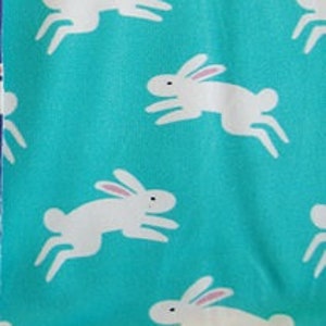 May include: A turquoise fabric with a repeating pattern of white rabbits in a running pose. The rabbits have pink inner ears and black eyes. The fabric is likely cotton or a similar material, suitable for quilting or crafting projects.