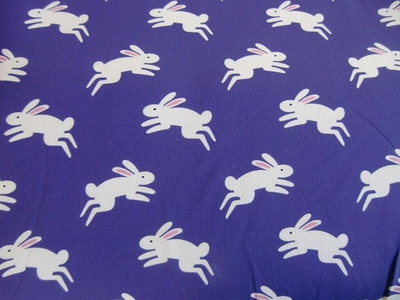 May include: A pattern of white rabbits with pink ears on a deep purple background. The rabbits are depicted in a running pose, creating a playful and whimsical design. This fabric is suitable for various craft projects.
