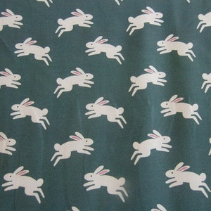 May include: A fabric with a repeating pattern of white rabbits with pink ears on a teal background. The rabbits are depicted in a running pose, creating a playful and whimsical design. This pattern is suitable for various craft projects.