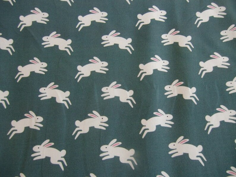 May include: A fabric with a repeating pattern of white rabbits with pink ears on a teal background. The rabbits are depicted in a running pose, creating a playful and whimsical design. This pattern is suitable for various craft projects.