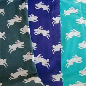 May include: Three fabric swatches in teal, navy, and green, each patterned with white rabbits in a running pose. The rabbits have pink ears. The fabric is likely cotton or a similar material, suitable for sewing projects.
