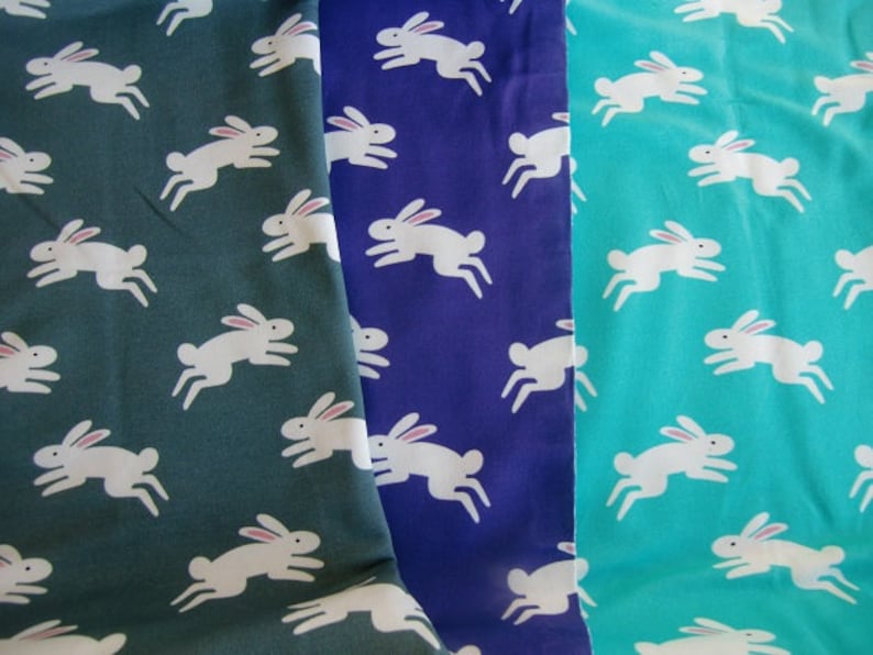 May include: Three fabric swatches in teal, navy, and green, each patterned with white rabbits in a running pose. The rabbits have pink ears. The fabric is likely cotton or a similar material, suitable for sewing projects.