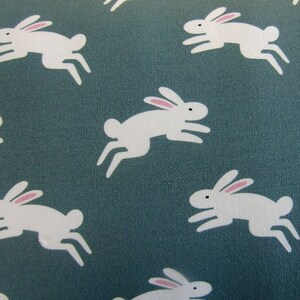 May include: A teal fabric with a repeating pattern of white rabbits in mid-leap. Each rabbit has pink inner ears and black eyes. The design is simple and playful, suitable for children's items or spring decor.