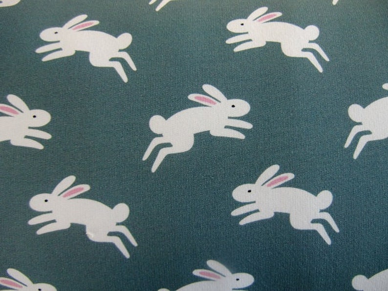 May include: A teal fabric with a repeating pattern of white rabbits in mid-leap. Each rabbit has pink inner ears and black eyes. The design is simple and playful, suitable for children's items or spring decor.