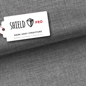 Pode incluir: Etiqueta branca com texto preto que diz "SHIELD PRO" e "DARK GREY STRUCTURE" com um símbolo de direitos autorais e o ano de 2020 seguido de "SUSANNE FIRMENICH". A etiqueta tem um cordão vermelho preso a ela e um logotipo de escudo preto e branco com uma árvore dentro.