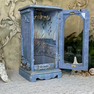 May include: A blue wooden cabinet with a hinged glass door, featuring a nautical-themed interior painting of seagulls, tall grass, and a sailboat. The exterior is decorated with relief carvings of a lighthouse and rope, with a metal hook rack inside.