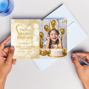 May include: Gold glitter birthday invitation with a photo of a girl smiling with a cake and balloons. The invitation says "Janie's Golden Birthday" and includes the date, time, and location of the party. RSVP information is also included.