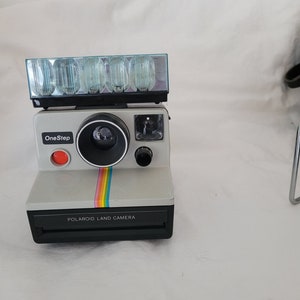 Peut inclure: Un appareil photo Polaroid OneStep Land Camera gris avec un flash intégré et une bande arc-en-ciel sur le devant.