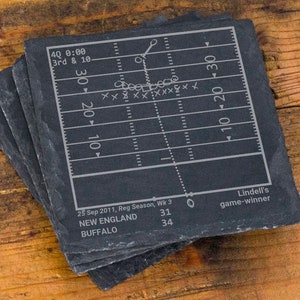 May include: A set of four black slate coasters featuring a football field with a game play diagram. The text on the coasters reads "4Q 0:00 3rd & 10", "25 Sep 2011, Reg Season, Wk 3", "NEW ENGLAND 31", "BUFFALO 34", "Lindell's game-winner".