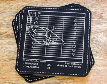 Peut inclure: Un ensemble de dessous-de-verre en cuir noir avec un motif de terrain de football blanc. Le motif comprend un schéma de jeu et le texte "10 11:28 4th & 5", "25 Nov 1971, Reg Season", "1 NEBRASKA 35", "2 OKLAHOMA 31", et "Punt Return in Game of the Century".