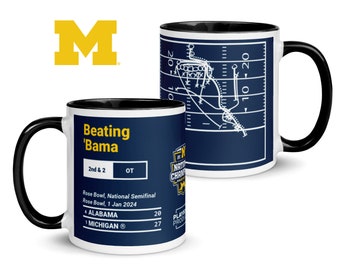 May include: A white ceramic mug with a black rim and handle. The mug features a blue and white illustration of a football play on one side and a blue and white scorecard on the other side. The scorecard shows the University of Michigan beating the University of Alabama 27-20 in the Rose Bowl National Semifinal on January 1, 2024. The text "Beating 'Bama" is printed on the mug.