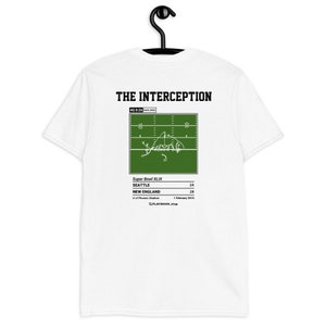 Peut inclure: T-shirt blanc avec un graphique vert représentant un schéma de jeu de football américain. Le graphique comprend le texte "THE INTERCEPTION", "4Q 0:26", "Super Bowl XLIX", "Seattle 24", "New England 28", "U of Phoenix Stadium", "1 February 2015", et "PLAYBOOK shop".
