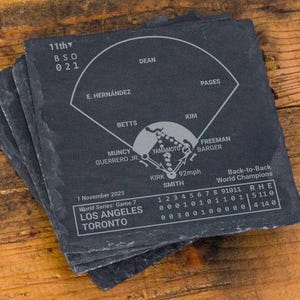 May include: A stack of four square slate coasters. Each coaster features a baseball field diagram with player names and game details. The coasters are dark gray with white etched details, resting on a wooden surface.