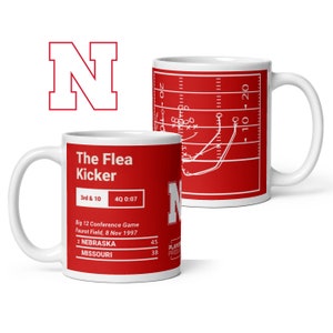 May include: White ceramic coffee mug featuring a red and white football play diagram. The mug displays the text "The Flea Kicker" and the score of a football game: "2 NEBRASKA 45, MISSOURI 38".