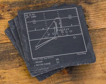May include: A square slate coaster featuring an engraved white graphic of a football play diagram. The text reads 4Q 0:45 2PT CONV, The Play, 15 Oct 1988, Reg Season, 1 MIAMI 30, 4 NOTRE DAME 31. The coaster has a dark gray, textured surface.