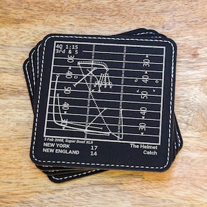 May include: Set of four black leather coasters with a white football field design. The coasters feature the play from Super Bowl XLII, with the score and date. The text "The Helmet Catch" is also included.