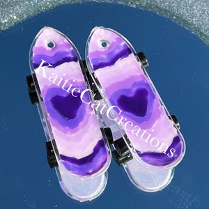 May include: Two miniature purple and white skateboard earrings with black wheels. The earrings are made of resin and have a heart shape in the center. The earrings are labeled "KaitleCatCreations".