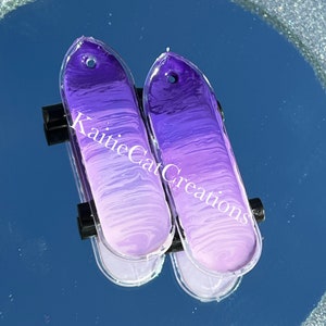 May include: Two purple and white ombre skateboard charm earrings. The charms are shaped like skateboards with a rounded top and a flat bottom. The charms are attached to silver earring hooks.