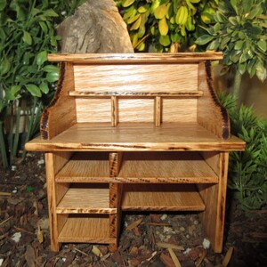 May include: A miniature wooden desk with a top shelf, a lower shelf with three compartments, and a bottom shelf with two compartments. The desk is made of light wood with a dark wood stain.