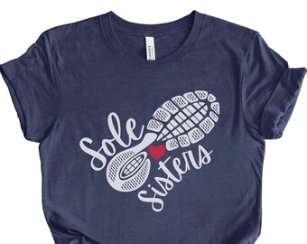 May include: Navy blue t-shirt with the text "Sole Sisters" in white script, featuring a white shoe sole design and a red heart. The shirt is made of a soft material.