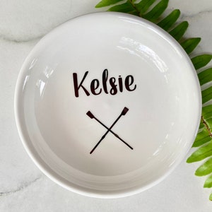 May include: White ceramic ring dish with the name "Kelsie" written in black cursive lettering and two black crossed oars.