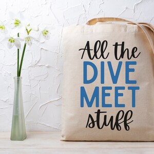 May include: A beige canvas tote bag with the text "All the DIVE MEET stuff" in black and blue. The bag has a simple design with long handles. A clear glass vase with white flowers is to the left of the bag.