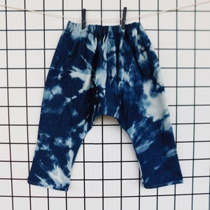 May include: Blue and white tie-dye pants hanging on a clothesline. The pants have a relaxed fit with an elastic waistband. The fabric is a dark blue with white cloud-like patterns.