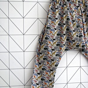 May include: A pair of patterned pants featuring an elastic waistband and a colorful animal print. The print includes elephants, giraffes, flamingos, and other animals on a white background. The pants are hanging on a clothesline with a wooden clothespin.