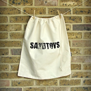 Puede incluir: Una bolsa de cordón de color crema con la palabra "SANDTOYS" impresa en negro, colgada de un tendedero con pinzas de madera contra una pared de ladrillos. La bolsa está hecha de una tela suave y natural.