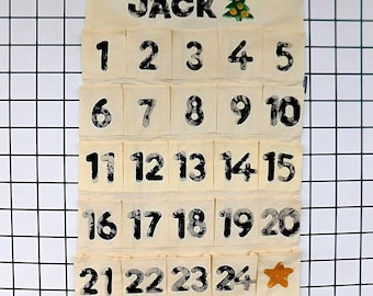 Op de afbeelding: Een crèmekleurige stoffen adventskalender die aan een houten stok met een touwtje hangt. Het heeft vijf rijen zakjes, elk bedrukt met zwarte cijfers van 1 tot 24. De naam JACK staat bovenaan met een kleine groene boom en een gouden ster.