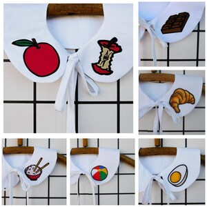 May include: White detachable collars with embroidered designs. The collars feature various food and object motifs, including an apple, apple core, chocolate bar, croissant, bowl of rice with chopsticks, beach ball, and a fried egg.