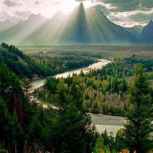 Puede incluir: Una vista panorámica de una cordillera con rayos de sol que brillan a través de las nubes. Un río serpentea a través de un valle de árboles y arbustos verdes. El río es visible en primer plano, y las montañas están en el fondo.