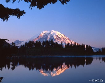 May include: A scenic landscape featuring a snow-capped mountain reflecting in a calm lake. The mountain is illuminated by the setting sun, with a forest of trees along the shoreline. The sky is a gradient of blue.