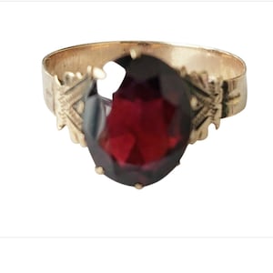 May include: A gold-toned ring featuring a large, faceted, teardrop-shaped, deep red gemstone. The band has decorative floral or leaf-like accents on each side of the gemstone. The ring is set against a white background.