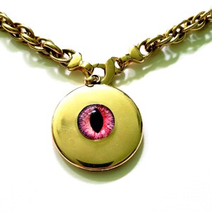 May include: A gold-toned necklace with a round pendant featuring a pink and black eye design. The chain is a thick, textured link, and the pendant has a polished finish. The eye has a detailed iris and pupil.