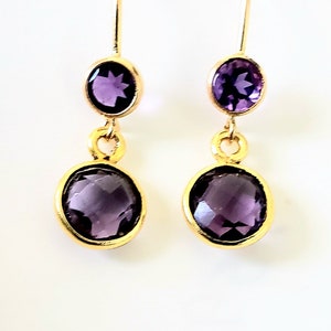 May include: Gold-tone drop earrings featuring two purple amethyst gemstones. The earrings have a simple design with a small round stone at the top and a larger round stone at the bottom, both set in gold bezels.
