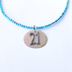 May include: A turquoise beaded necklace with a gold pendant featuring the number "21" in a silver font. The pendant is oval-shaped and has a gold bail. The necklace is on a white background.