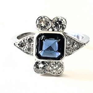 May include: A silver ring featuring a square, blue gemstone at the center, flanked by two pairs of round, clear gemstones. Additional small, clear gemstones are set on either side of the central stone, creating a symmetrical design. The ring has a classic, elegant style.